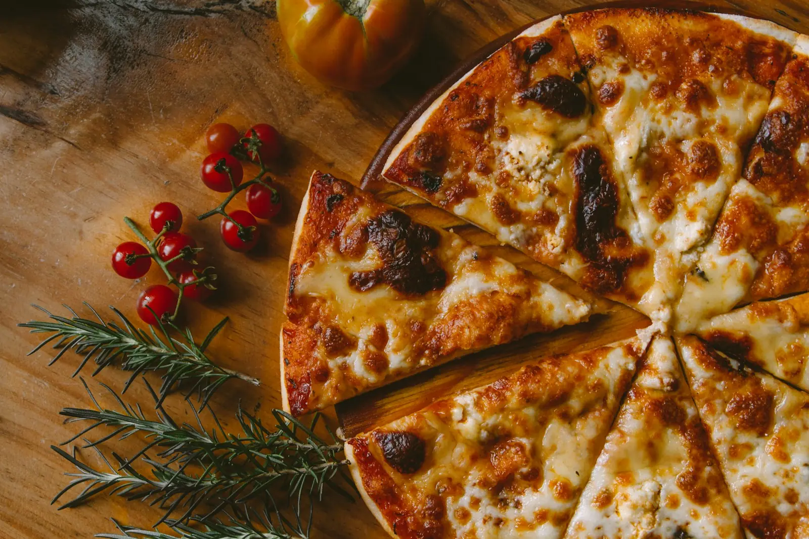Pizza fatiada sobre uma mesa de madeira, com tomates-cereja e ramos de alecrim